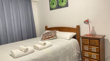 Un lit bien fait, avec du linge de lit blanc et deux serviettes enroulées, une tête de lit en bois, un coussin à motifs, une petite commode en bois avec des tiroirs et une lampe de bureau rouge.