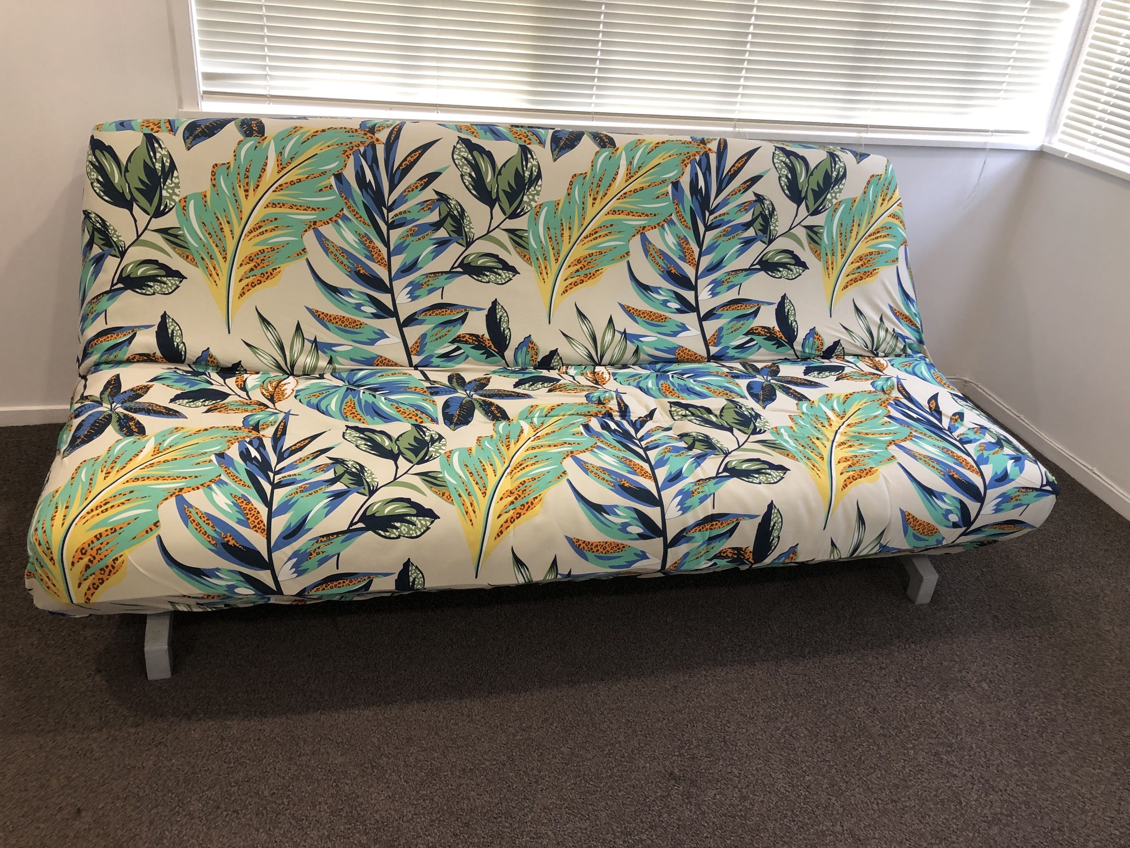 A sofa with a floral pattern, positioned in a room with a carpeted floor and a window with blinds.