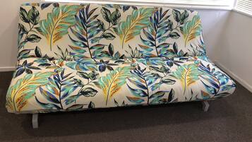 A sofa with a floral pattern, positioned in a room with a carpeted floor and a window with blinds.
