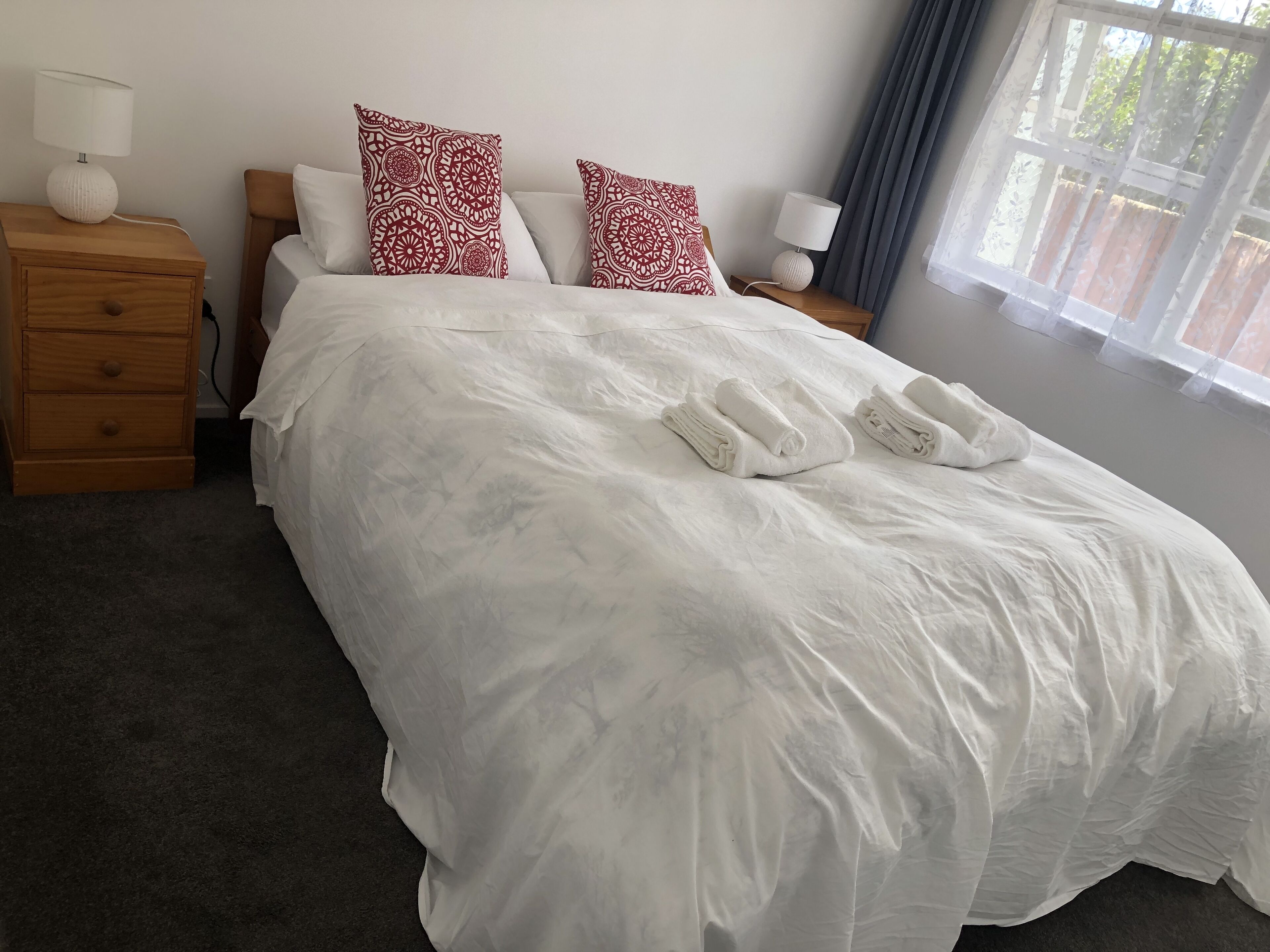 Una cama bien tendida con sábanas blancas y dos almohadones decorativos, una mesita de noche de madera con una lámpara, y una ventana con cortinas.