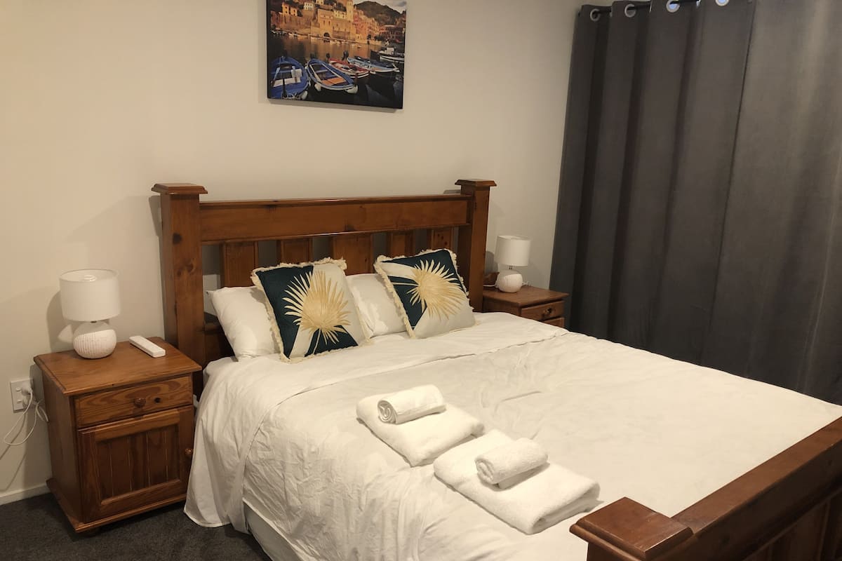 Cama bem arrumada com lençóis brancos, duas mesas de cabeceira com abajures e um quadro de uma paisagem costeira na parede.