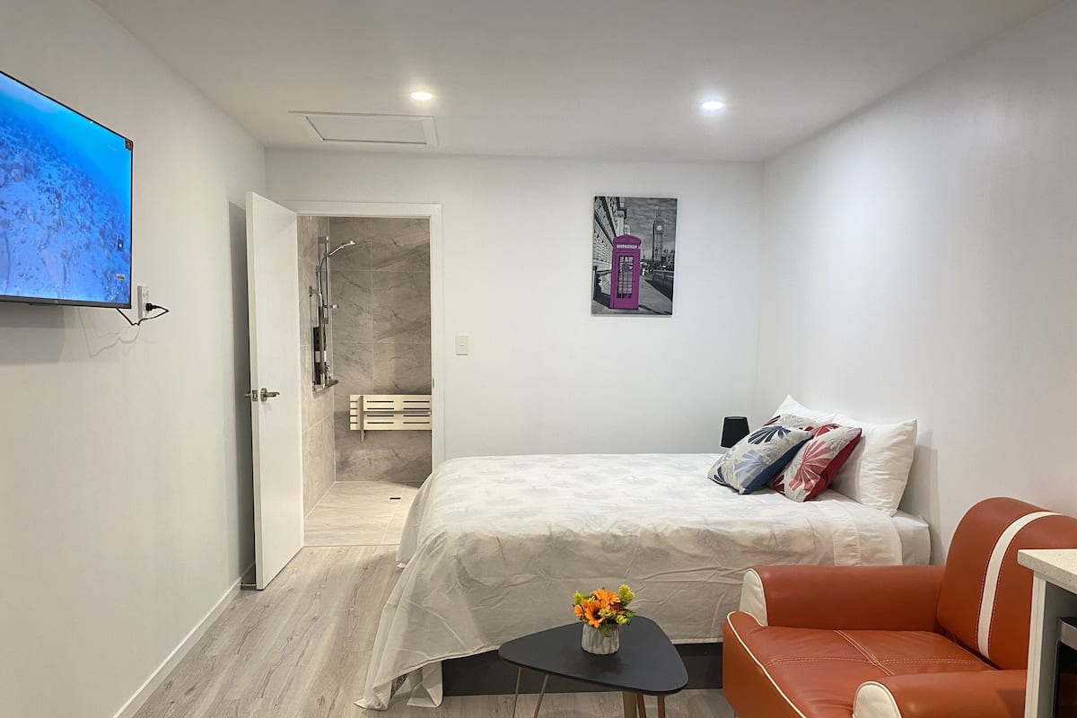 Um quarto moderno com uma cama, uma mesinha com flores, um sofá de couro vermelho e uma televisão na parede.