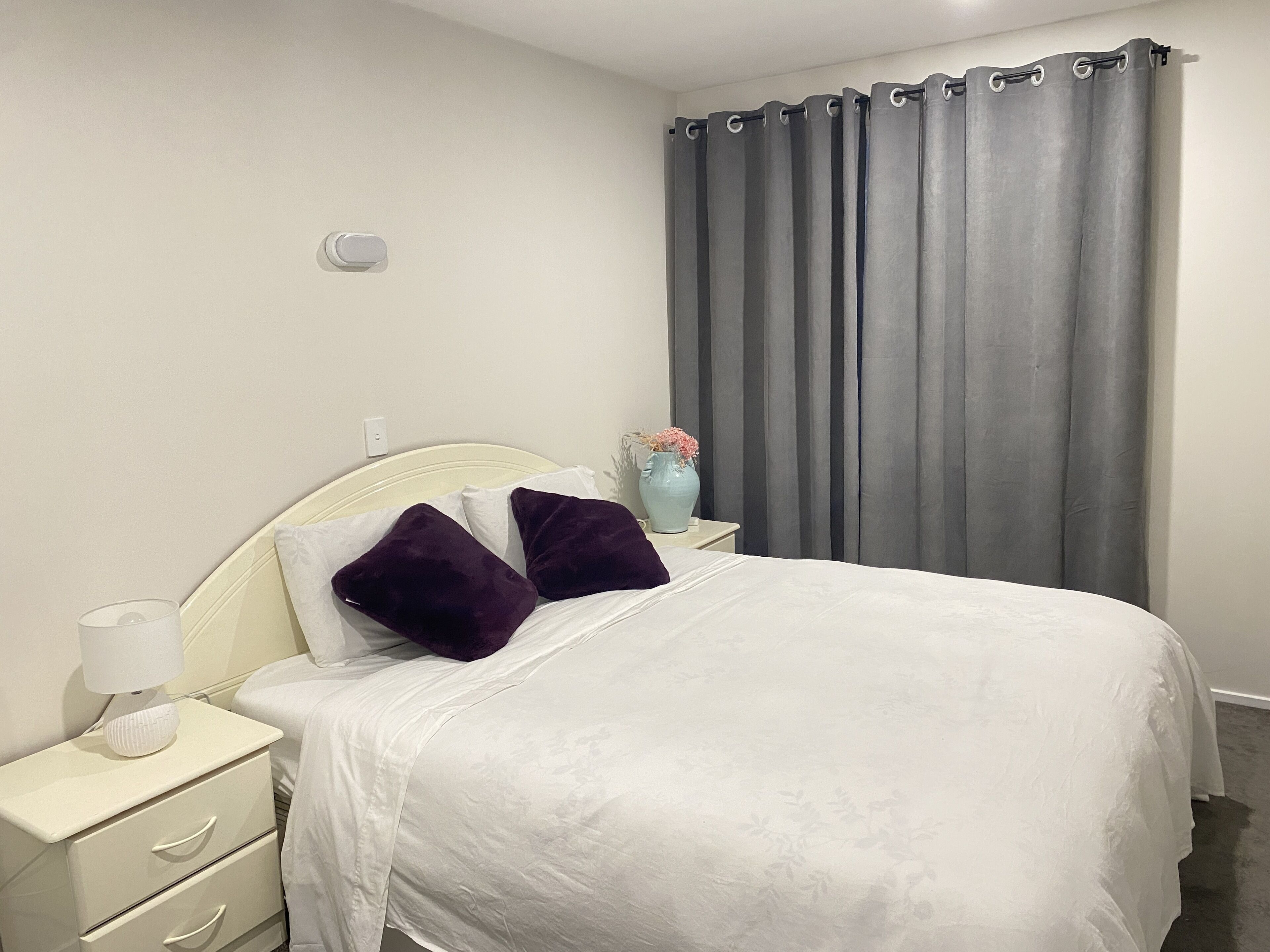Un dormitorio con cama, mesita de noche blanca y un florero con flores.