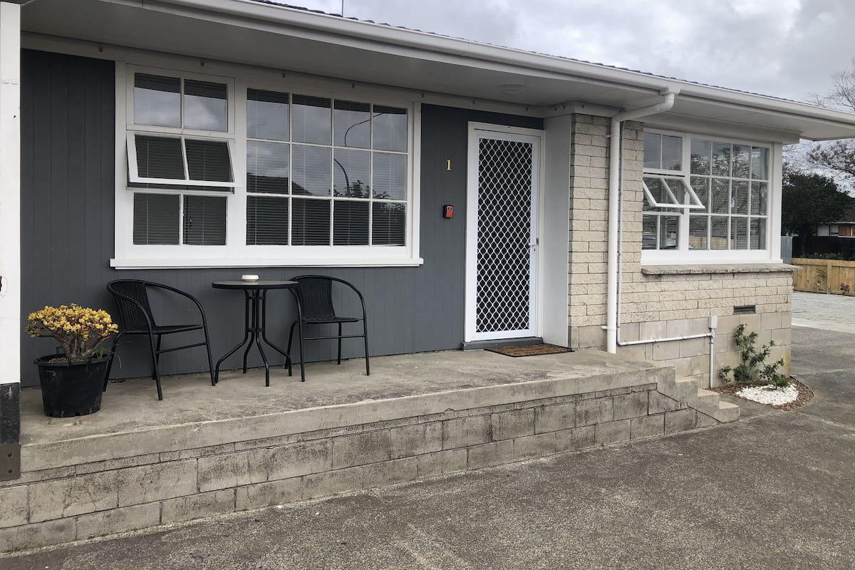 Um edifício residencial com fachada cinzenta, janelas emolduradas por branco, porta com grade metálica e uma pequena área externa com duas cadeiras e uma mesa.