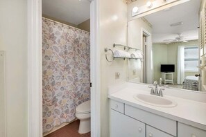 Une salle de bain avec un rideau de douche à motifs floraux, des toilettes blanches, un lavabo avec un miroir et un ventilateur de plafond.