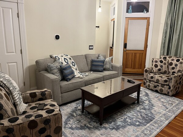 Un salon avec un canapé gris, des fauteuils à motifs, une table basse en bois et un tapis à motifs.