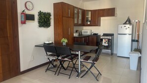 Uma cozinha compacta com armários de madeira, micro-ondas, fogão e geladeira. Há uma mesa de jantar com cadeiras e um relógio na parede.