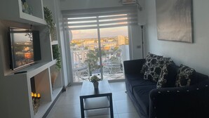 Uma sala de estar com uma janela grande, uma TV de tela plana, um sofá com almofadas estampadas e uma mesinha com uma planta.