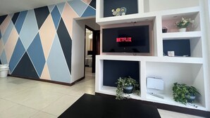 Uma sala de estar moderna com uma televisão de tela plana grande exibindo 'NETFLIX', uma parede com padrão geométrico e um vaso de planta.