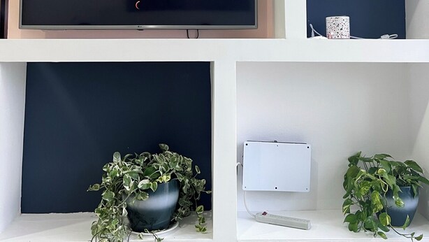 Uma sala de estar moderna com uma grande TV de tela plana exibindo o logotipo da Netflix, cercada por estantes brancas com objetos decorativos e plantas.