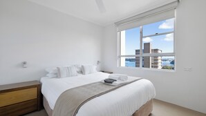 Un dormitorio con una cama grande, una mesita de noche de madera, un ventilador de techo y una ventana con vista a la ciudad.