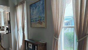 A room with wooden flooring, a cabinet with colorful drawers, a painting of a beach scene, and a window with curtains. - Anchors Aweigh (Cape Charles)