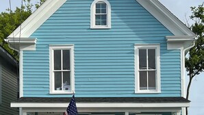 A two-story house with a blue exterior and white trim, featuring a front porch with two chairs and an American flag. - Anchors Aweigh (Cape Charles)
