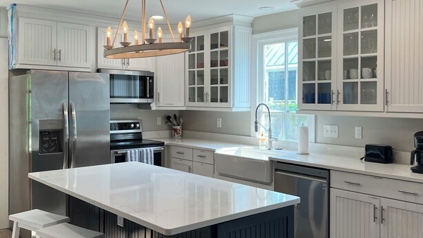 A modern kitchen with white cabinetry, a central island with stools, stainless steel appliances, and a pendant light fixture. - Anchors Aweigh (Cape Charles)