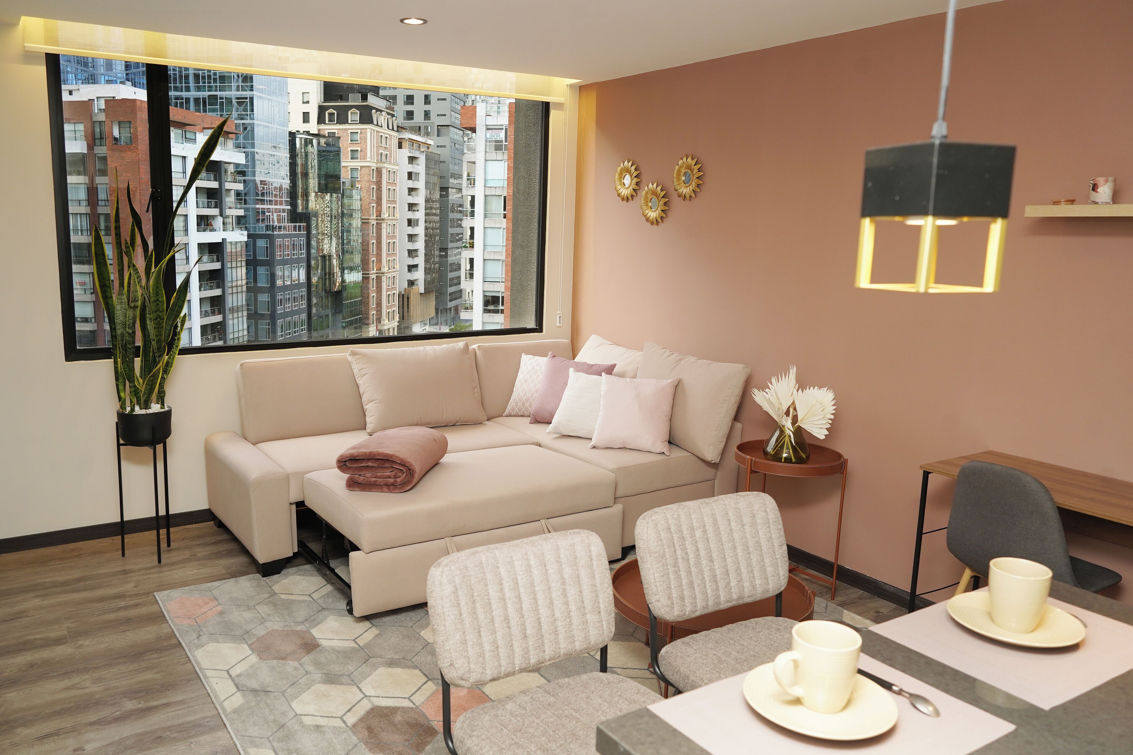 Uma sala de estar moderna com sofá, cadeiras, mesa de jantar com xícara e pires, planta em vaso e vista da cidade por grandes janelas.
