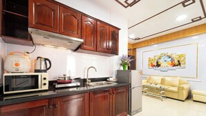 A kitchen with wooden cabinets, a microwave, a kettle, and a refrigerator. There is a sink and a vase with flowers on the counter. A beige sofa and a coffee table are in the living area.