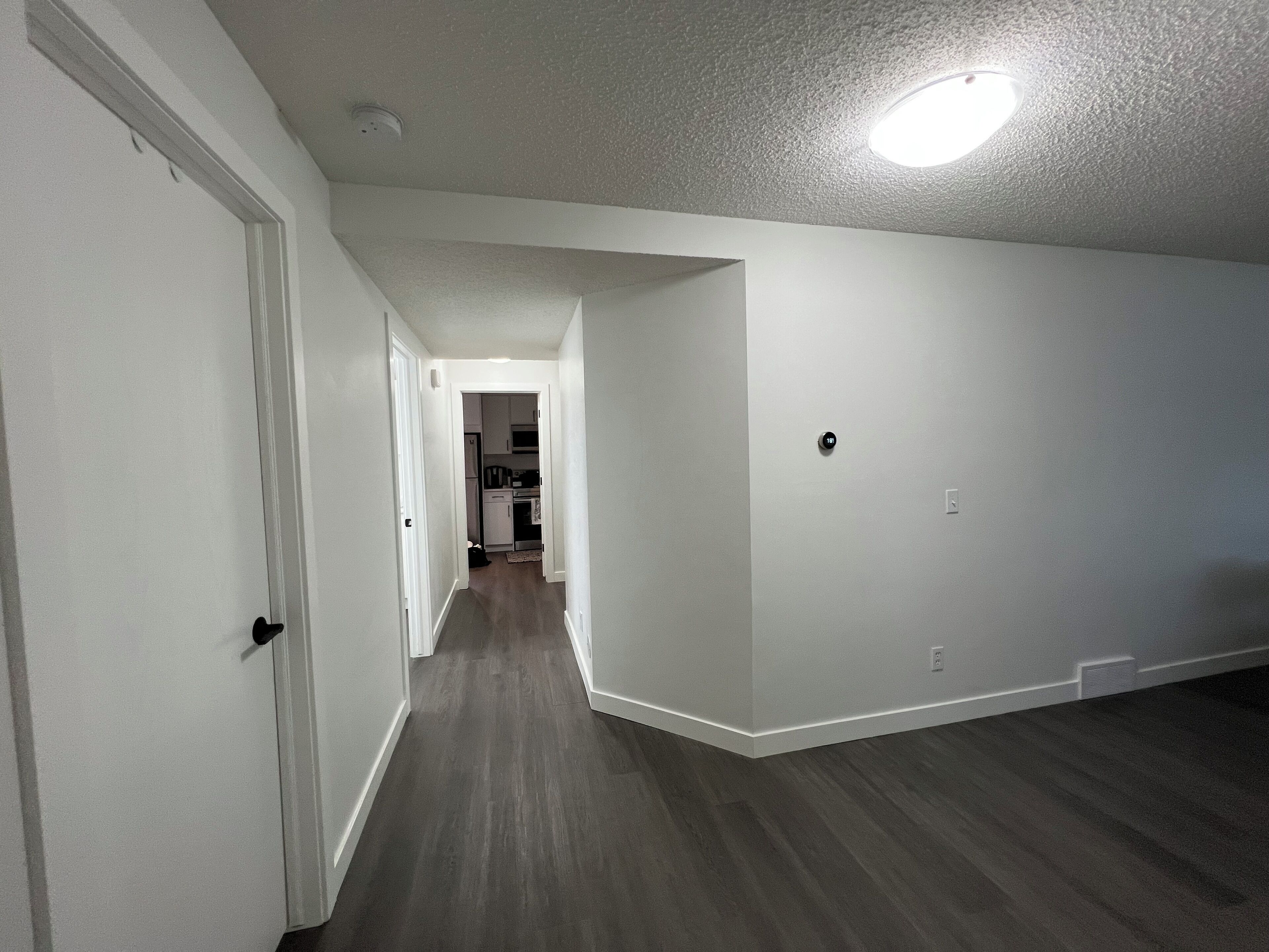 A hallway with white walls and dark wooden flooring, leading to a kitchen area with cabinets and appliances.