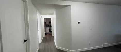 A hallway with white walls and dark wooden flooring, leading to a kitchen area with cabinets and appliances.