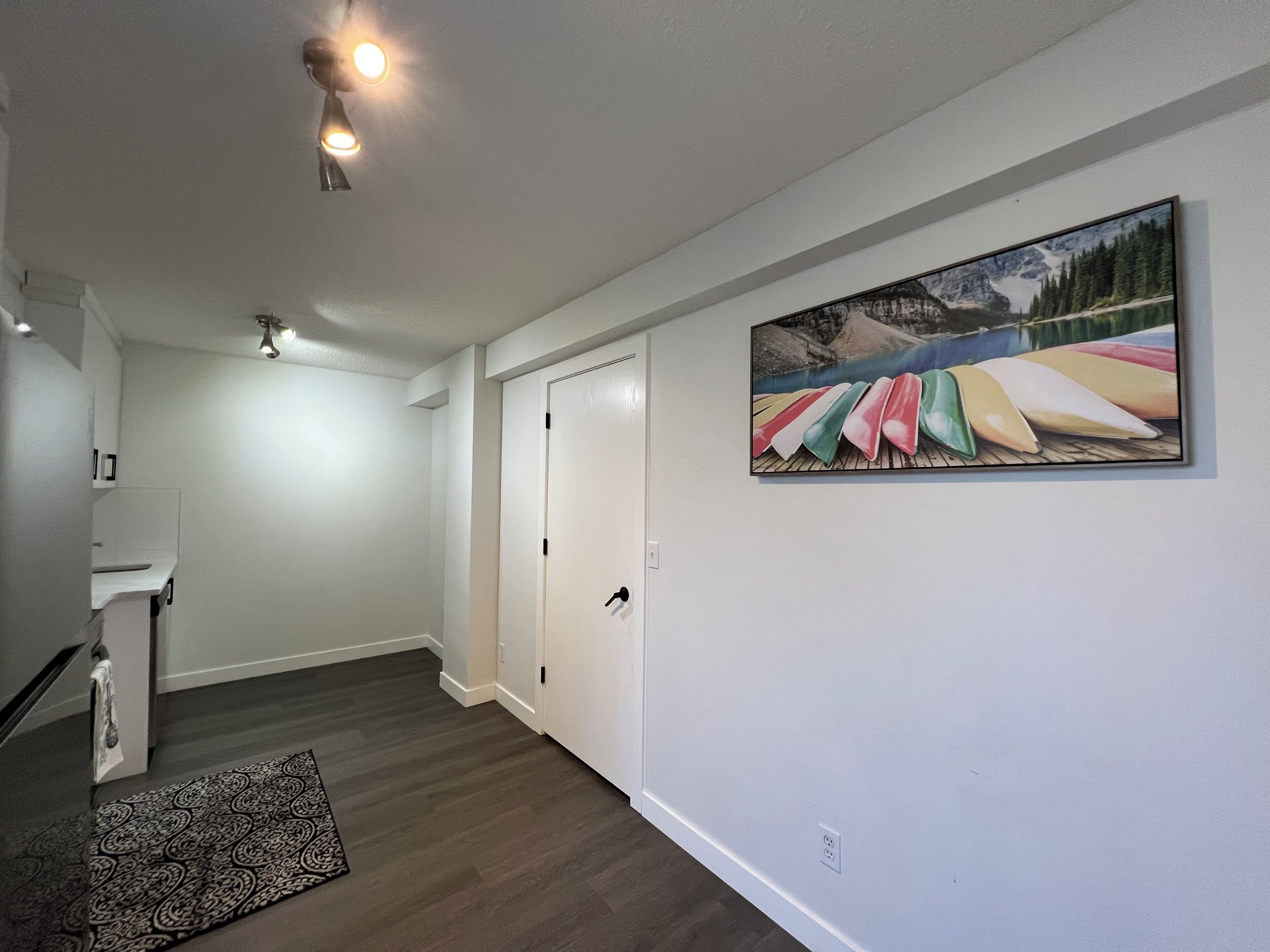 A hallway with a wooden floor, a patterned rug, a door, and a painting of colorful boats on a dock.