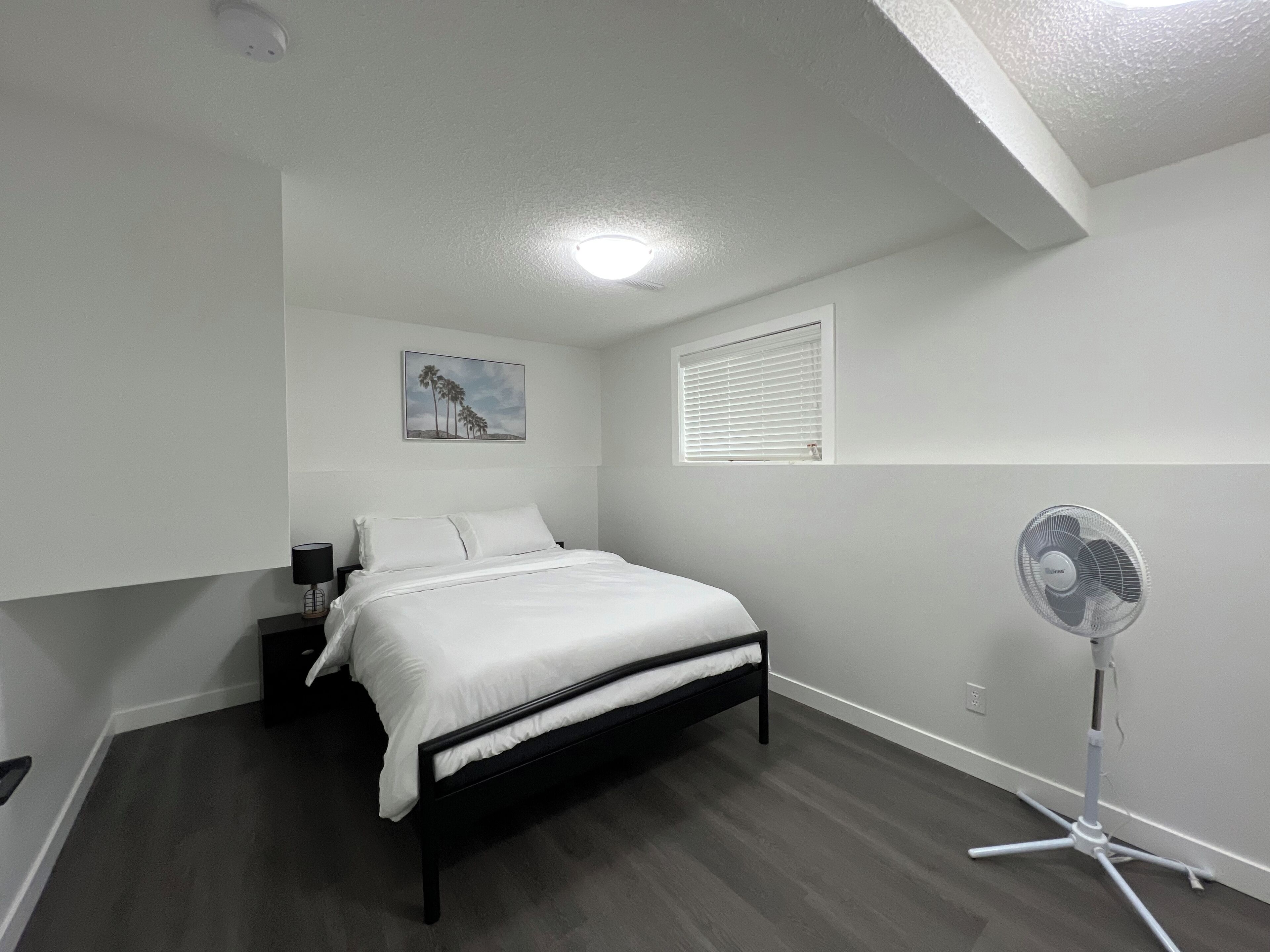 Een slaapkamer met een bed, een nachtkastje, een lamp, een aan de muur hangend schilderij, een raam met jaloezieën en een staande ventilator.