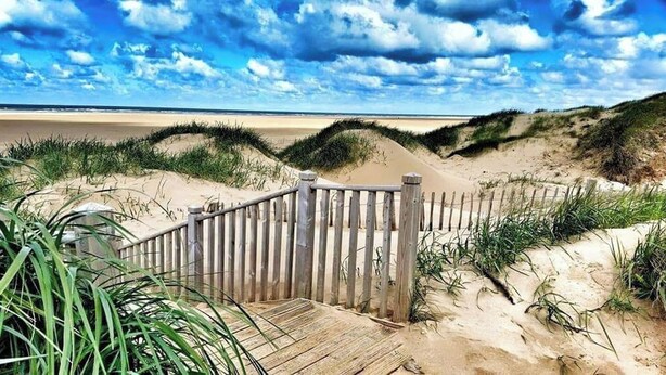 Un paseo marítimo de madera que conduce a una playa con dunas y cielo nublado.