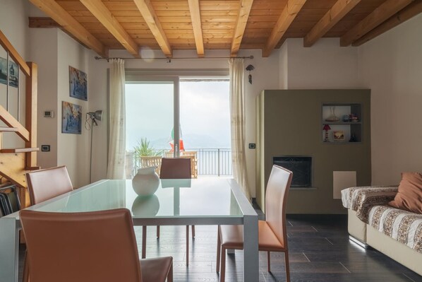 A modern living room with a glass dining table, leather chairs, a sofa, and a fireplace. There is a balcony with a view of the mountains.