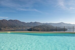 A swimming pool with clear blue water, surrounded by a metal fence, and a mountainous landscape in the background.