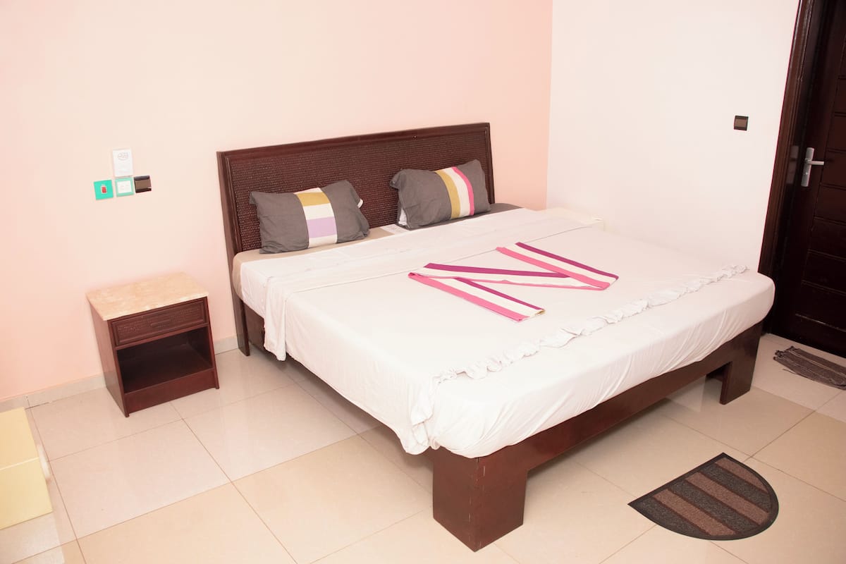 A neatly made bed with a white bedspread and pillows, a wooden headboard, a nightstand with a drawer, and a door to the right.