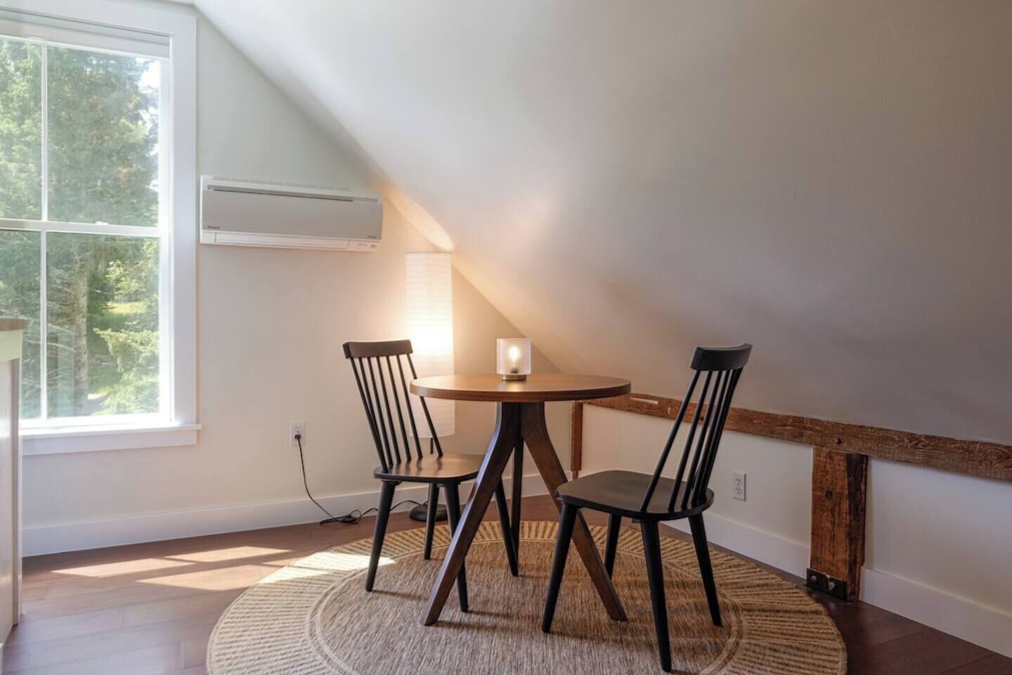 A dining area with a round table, two chairs, a candle, and a view of trees through a large window.