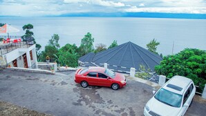 Un auto rojo estacionado en una entrada de casa, con una camioneta blanca cerca, con vista a una gran masa de agua y un paisaje montañoso.