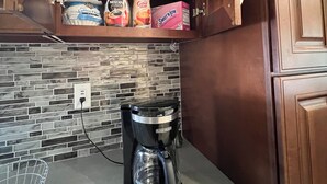 Een keuken met een koffiezetapparaat, een kast met koffieproducten en een betegelde spatwand.