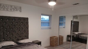 Un dormitorio con cabecera capitonada, cama con ropa de cama gris, una mesita de noche pequeña, un baúl de mimbre, una ventana con persianas y un espejo que refleja la cama y la ventana.