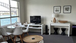 Un salon moderne comprenant un canapé, une table basse, un téléviseur et un bureau. Une fenêtre offre une vue sur un pont.