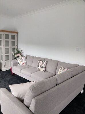 Ein graues Ecksofa mit floralen Kissen, eine weiße Vitrine mit Glastüren und ein dunkler Teppich.