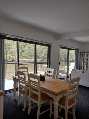 Ein Essbereich mit Tisch und Stühlen, große Fenster mit Blick auf Grünflächen und Wasser, und ein weißes Sofa im Hintergrund.