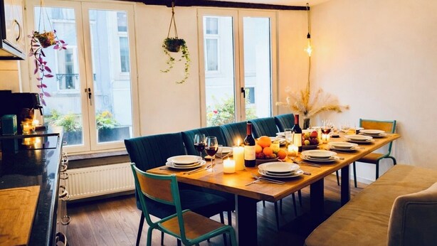 Un coin repas avec une table en bois dressée pour quatre personnes, entourée de chaises, et offrant une vue sur l’extérieur grâce à de grandes fenêtres.