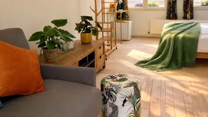 Une pièce chaleureuse avec un canapé, une table d’appoint en bois agrémentée de plantes, une échelle, un lit recouvert d’une couverture verte et une fenêtre avec des rideaux.