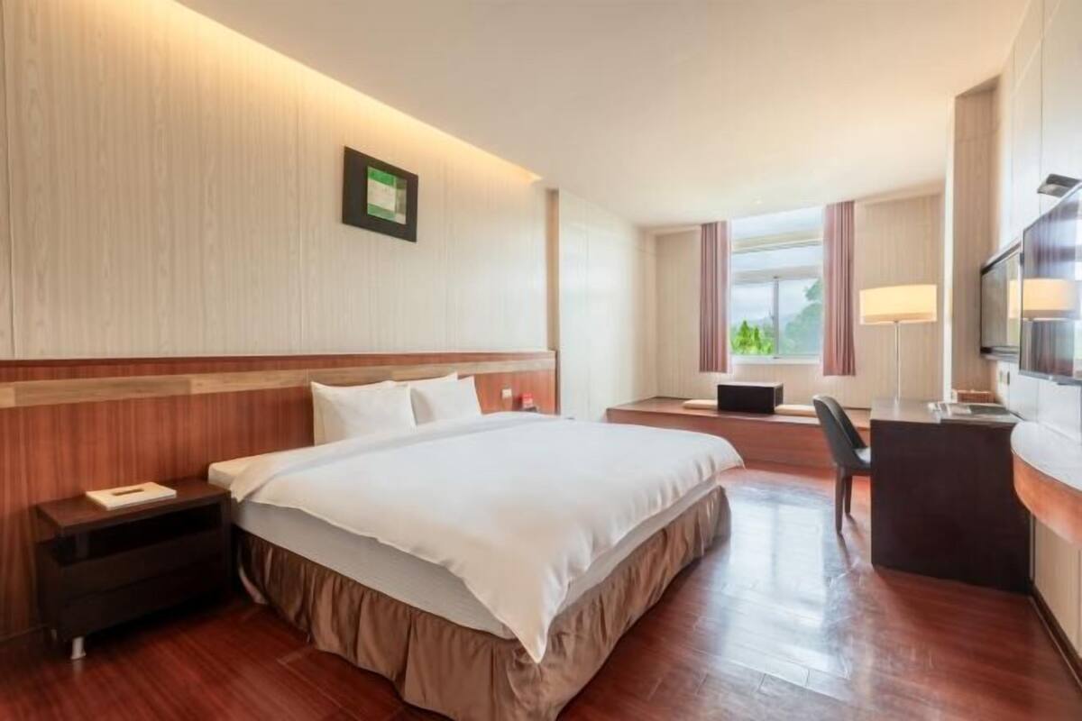 A hotel room with a bed, bedside table, desk, chair, TV, and a window with curtains.
