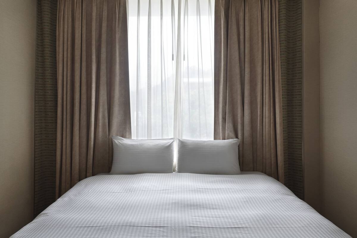 A neatly made bed with a white bedspread and two pillows, flanked by sheer curtains and heavier drapes, with a view of a cityscape through the window.