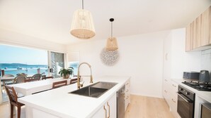 Une cuisine moderne dotée d’un grand îlot central, de meubles blancs et offrant une vue sur la mer grâce à de grandes fenêtres.