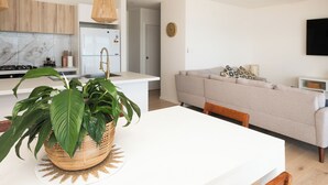 Une cuisine moderne avec un plan de travail blanc, une cuisinière encastrée et un réfrigérateur. On peut voir un panier en osier contenant des feuilles vertes sur le plan de travail.