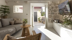 Una sala de estar moderna con un sofá, una mesa de centro y un televisor montado en una pared de piedra. Hay una puerta que da a un área exterior con plantas y una silla.