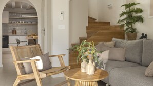 Una sala moderna con un sofá gris, una mesa de centro de madera y una silla de mimbre. Una escalera conduce a un nivel superior, y se ve una cocina al fondo.