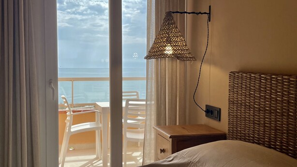 Une chambre avec un lit, une tête de lit en osier, une table de chevet, une lampe et une vue sur la mer depuis une porte-fenêtre donnant sur un balcon.