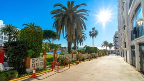 A sunny street with palm trees, a pool, and a building with balconies.