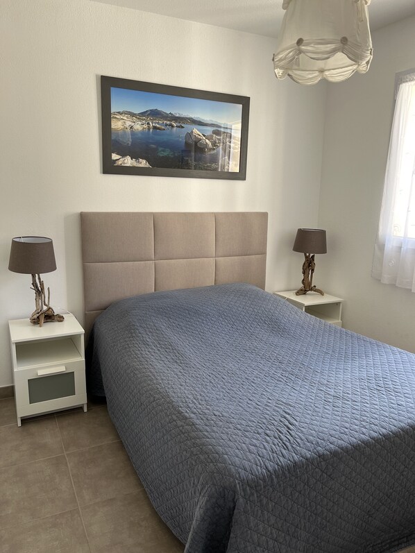 Een slaapkamer met een groot bed, een nachtkastje, een lamp, een ingelijst schilderij aan de muur en een raam met gordijnen.