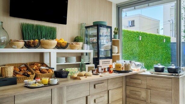 A modern kitchen with wooden cabinets, a glass display case, and a view of a green hedge outside. - B&B HOTEL Antibes Juan-les-Pins (Antibes)