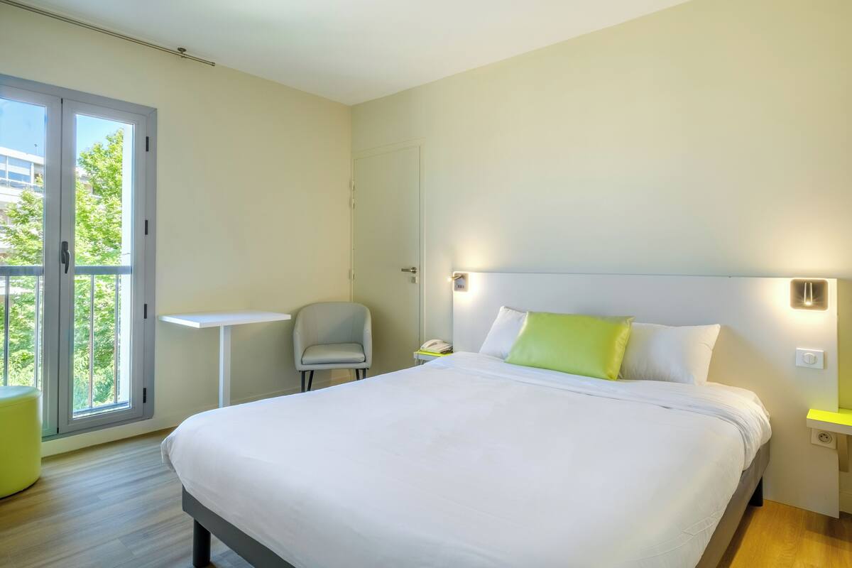 A modern hotel room with a large bed, a small desk, a chair, and a balcony with a view of buildings and trees.