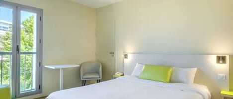 A modern hotel room with a large bed, a small desk, a chair, and a balcony with a view of buildings and trees.