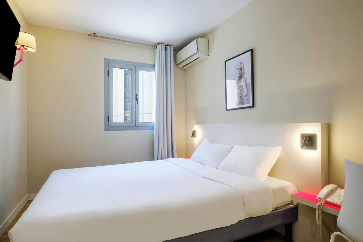 A hotel room with a large bed, a bedside telephone, a window with curtains, an air conditioning unit, and a framed picture on the wall.